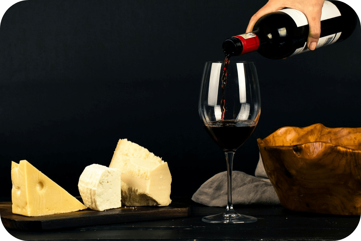 A glass of red wine being filled, with cheese on the table next to it.